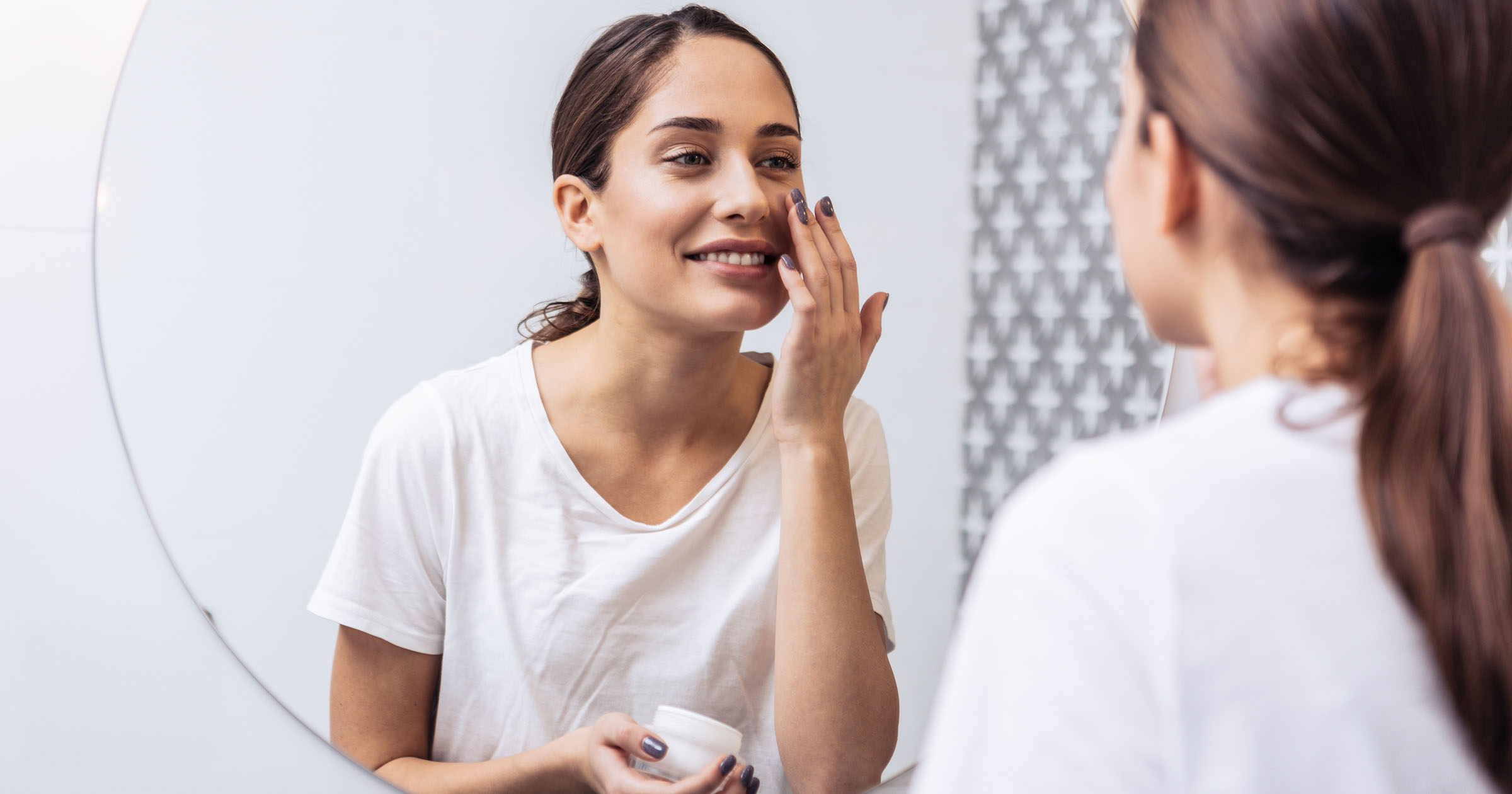 Ayurvedic Skincare: Adult woman applying moisturizer on her face, smiling at herself in the bathroom mirror, wearing a white T-shirt.