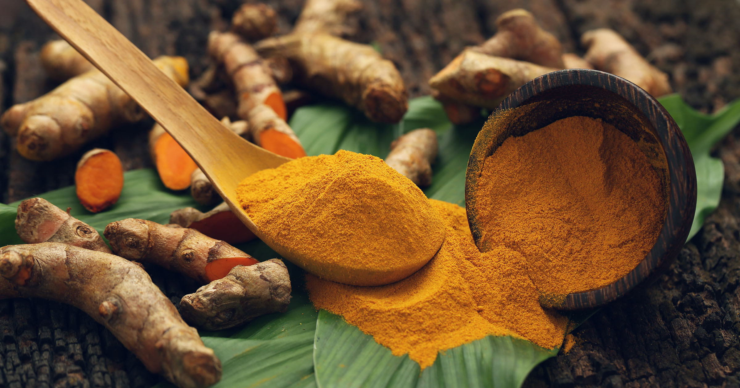 Turmeric Benefits: Turmeric powder on a wooden spoon with a leaf and roots in the background